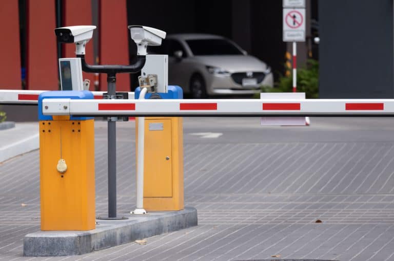 A barrier arm lowered across a paved vehicle entrance with a car waiting beyond it, a typical access point for car motion sensors.